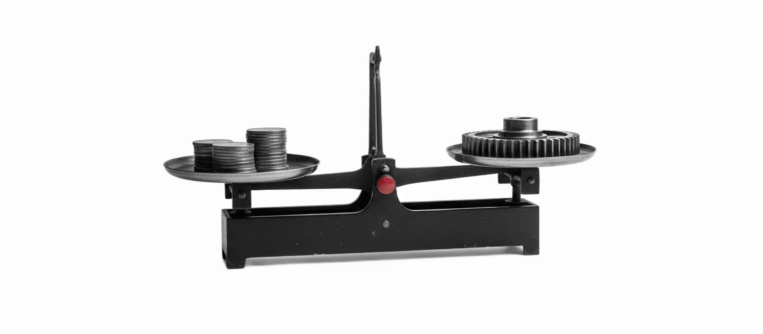 A black-and-white photograph of a 1970s mechanical balance scale weighing a stack of coins against a single brass gear — the cost-versus-system trade-off at the heart of the AI agency ROI comparison.