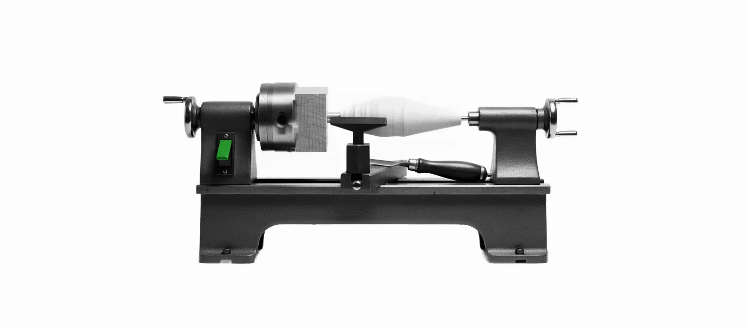 A wood lathe with a blank half-turned from a rough square block into a smooth spindle, a chisel resting on the tool rest, a green start switch, black and white.