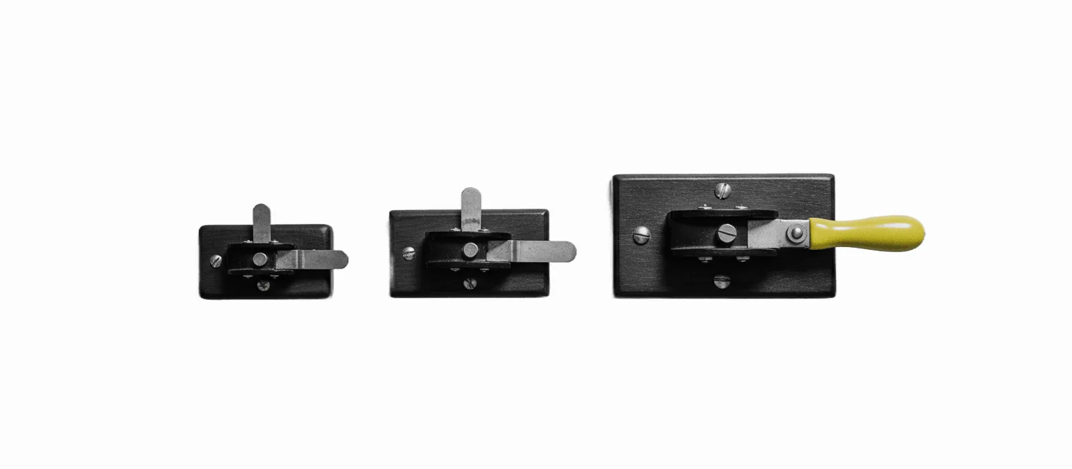 Three vintage electrical knife switches of increasing size mounted in a row, the largest with a single vivid yellow handle, on a white background